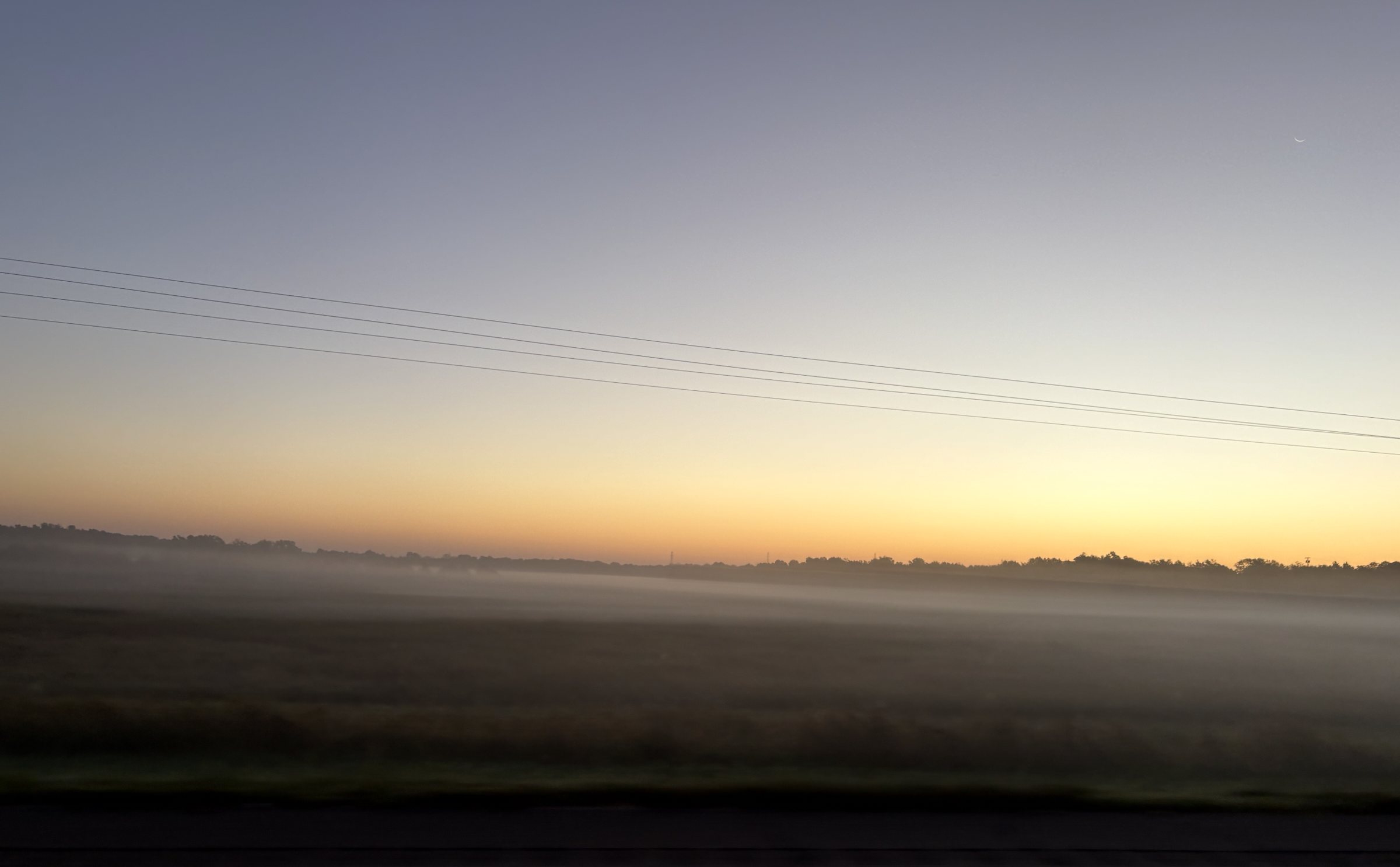 A field at first light: fog over the grass, power lines crossing toward an amber horizon, a thin moon above.